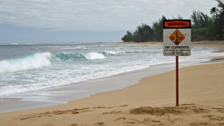 Una playa tropical de arena con una señal de corrientes y una ola rompiendo en el fondo. El cartel dice: "Rip Currents - You could be swept out and drown - If in doubt, don't go out".