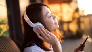 Retrato de una joven asiática sonriente con los ojos cerrados disfrutando de la música con auriculares y usando un teléfono inteligente, relajándose en el balcón contra la luz del sol.