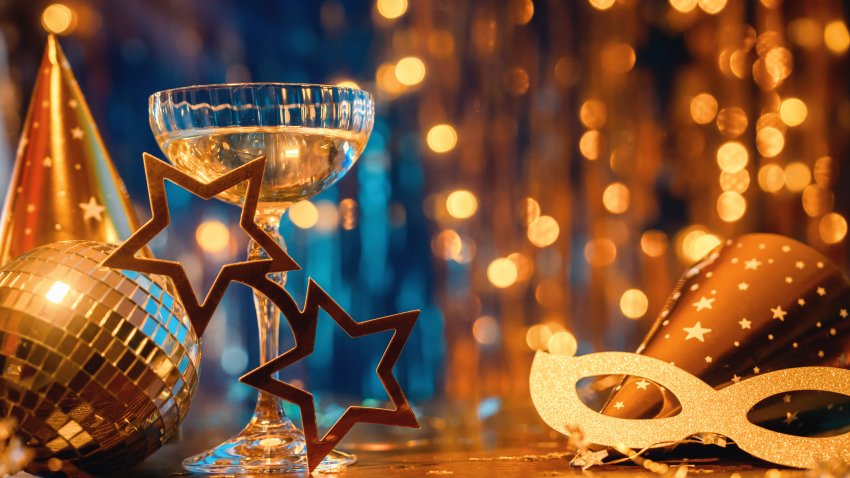 New Year or Christmas celebration concept with golden lights with disco ball and tinsel and Christmas tree with masquerade mask, fancy dress party hats and confetti. Champagne flute